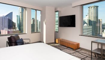 The image shows a modern hotel room with large windows, a bed, a chair, a wall-mounted TV, a wooden cabinet, and a cityscape view.