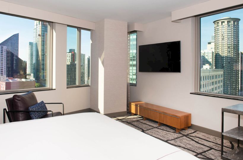 The image shows a modern hotel room with large windows, a bed, a chair, a wall-mounted TV, a wooden cabinet, and a cityscape view.