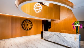 A modern lobby with a marble reception desk, spiral wooden architecture, wall clock, and a flower vase on the desk.