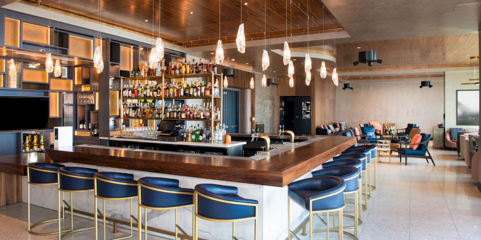 The image shows a modern bar with blue chairs, a wooden countertop, a well-stocked liquor shelf, and pendant lighting in a spacious, elegant room.