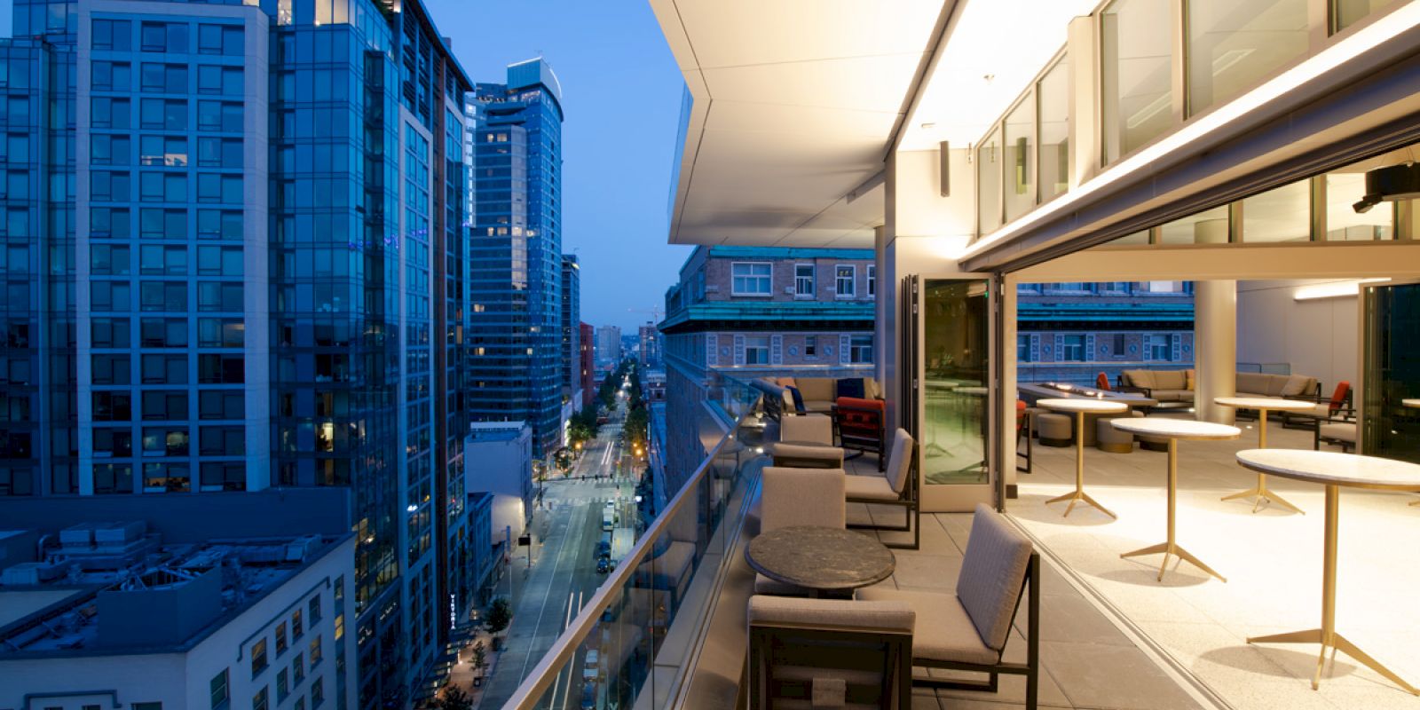 An elegant rooftop balcony with tables and chairs overlooks a cityscape with modern high-rise buildings during dusk.