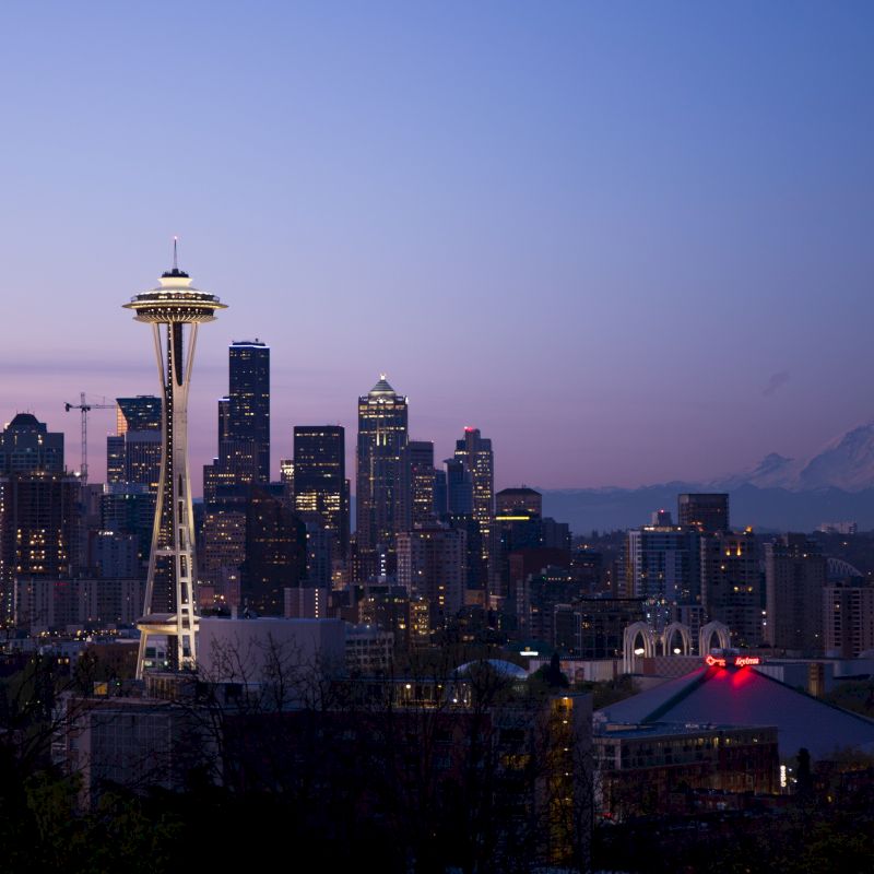 The image shows the Seattle skyline at dusk, featuring the Space Needle prominently, with Mount Rainier in the background and city lights beginning to illuminate.