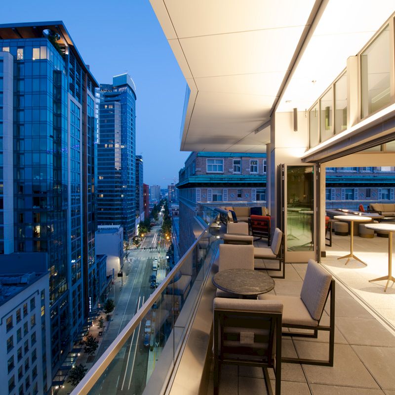 A rooftop patio with tables and chairs overlooks a cityscape with modern high-rise buildings during the evening.