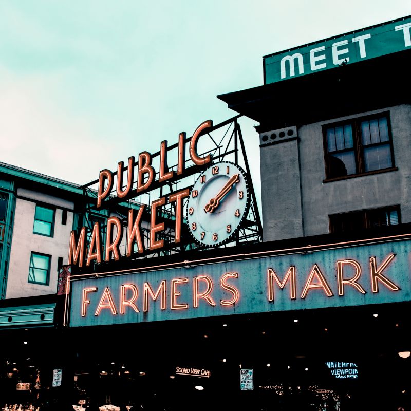 Image of an iconic public farmers market sign with a clock, under a cloudy sky, and with surrounding buildings, evoking a classic bustling market scene.