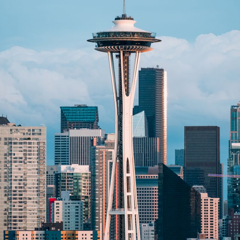 The image shows a cityscape with the Space Needle, surrounded by modern buildings and skyscrapers, set against a backdrop of partly cloudy sky.