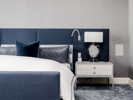A modern bedroom with a navy headboard, white bedding, side table with a lamp, and decorative object, against a grey wall, with a blue-grey carpet.