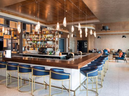 This image shows a modern bar with a wooden counter, blue barstools, a variety of bottles on display, stylish lighting, and seating areas.