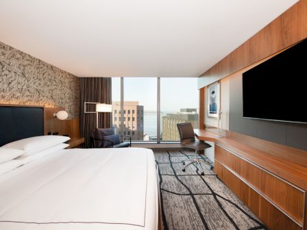 A modern hotel room with a large bed, desk, chair, TV, and window overlooking a cityscape and water.