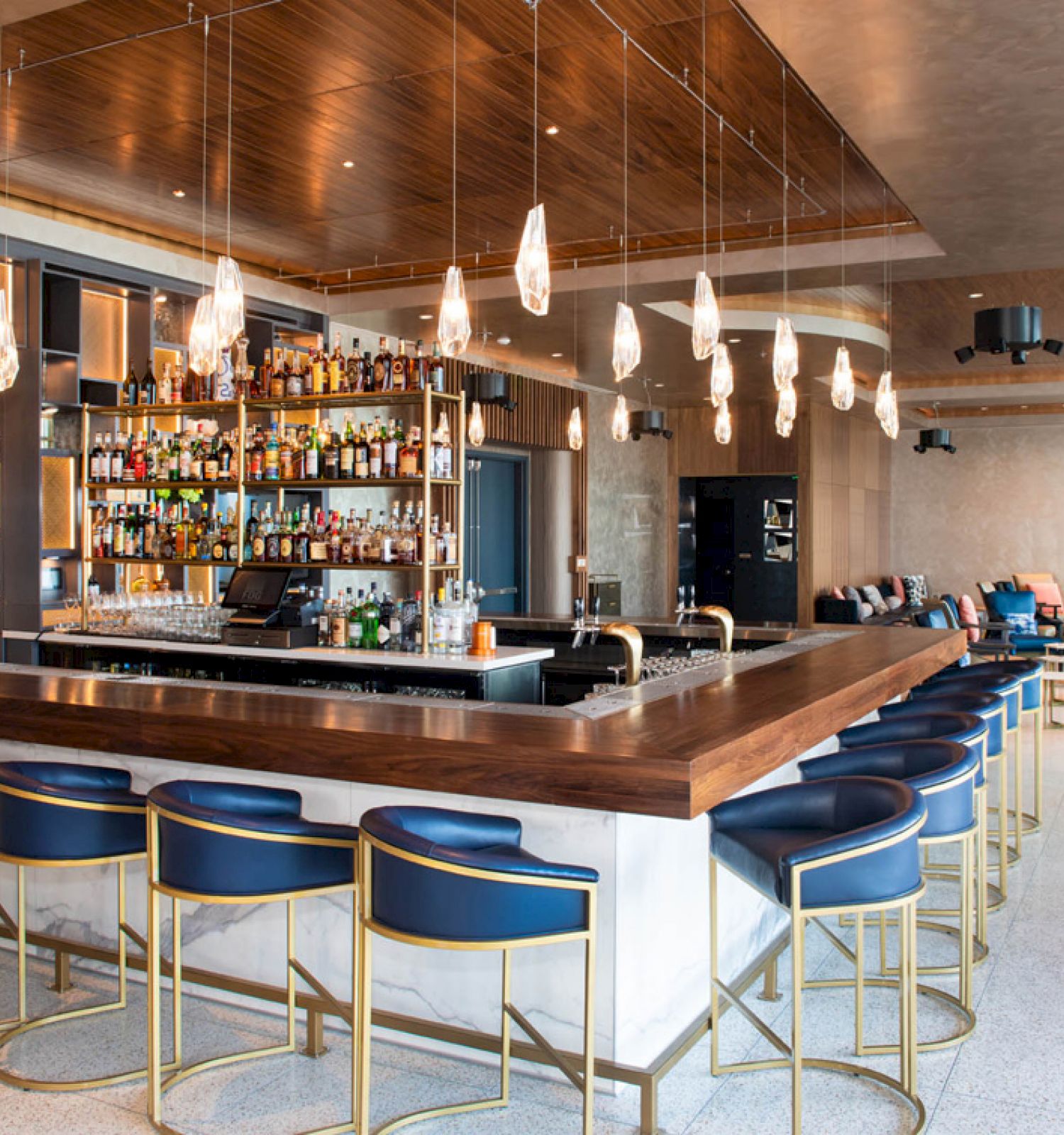 A modern bar with blue and gold stools, a fully stocked bar, hanging pendant lights, and a cozy seating area with couches in the background.