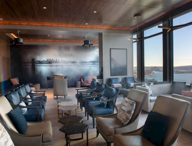 A modern lounge area with comfortable seating, large windows offering a water view, and stylish decor, including a wall mural depicting a cityscape.