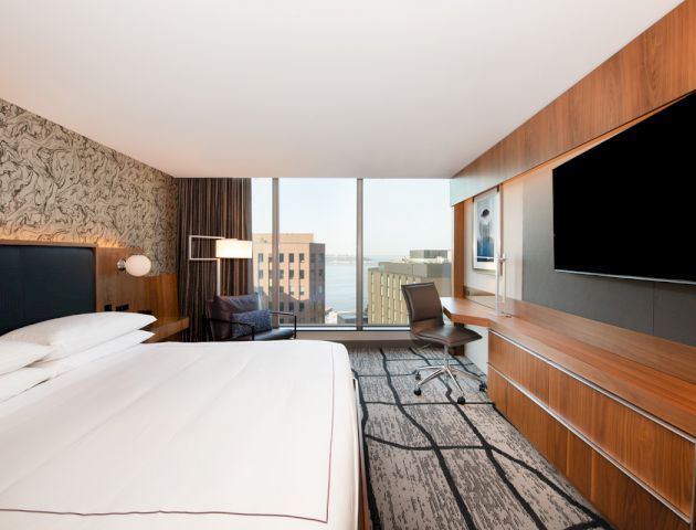 A modern hotel room with a bed, desk, chair, large window view of buildings, and a flat-screen TV mounted on the wall.