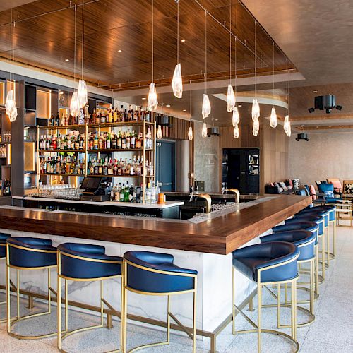 A modern bar with a wooden countertop, blue upholstered stools, hanging pendant lights, and a back wall filled with various bottles.