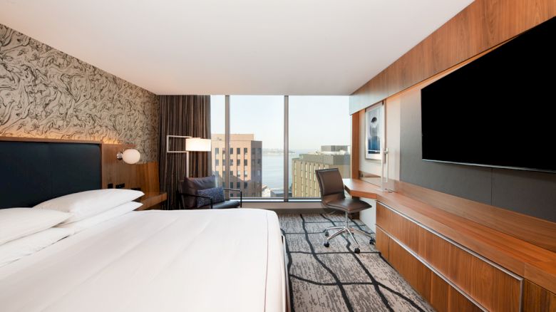 A modern hotel room features a large bed, flat-screen TV, desk with chair, and large window with a cityscape view and patterned accent wall.