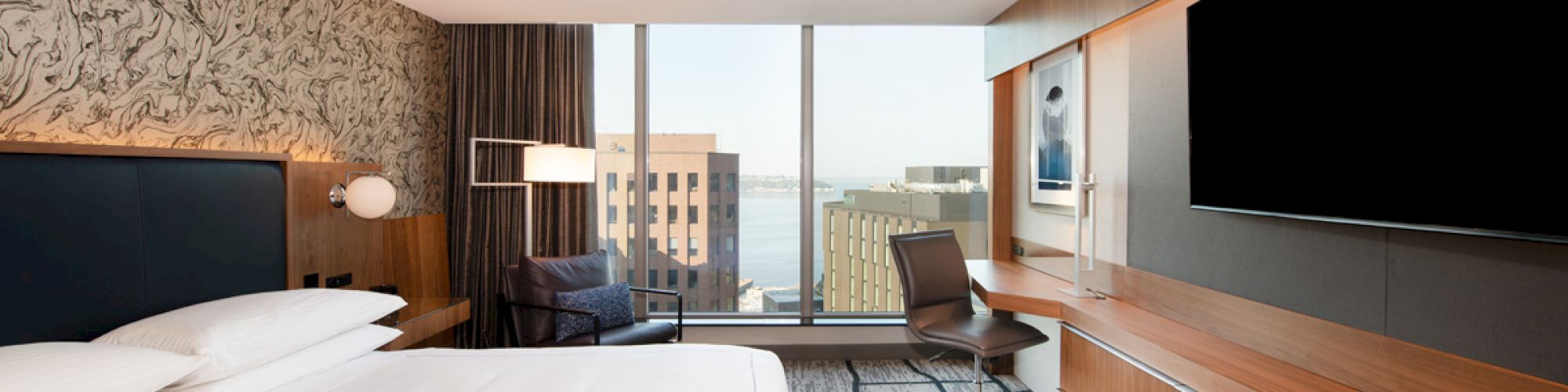 A modern hotel room with a large bed, a desk, a TV, and a view of buildings through floor-to-ceiling windows.